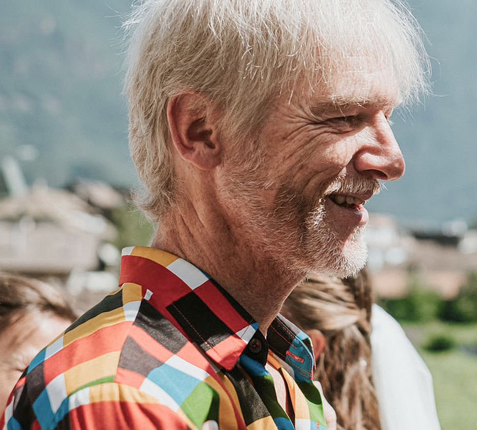 Uomo con capelli bianchi in camicia a quadri colorata che ride di profilo davanti a panorama montano